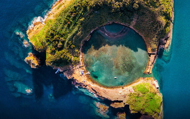 An aerial image of a mountainous green island with a water-filled crater in the center surrounded by deep blue ocean waters