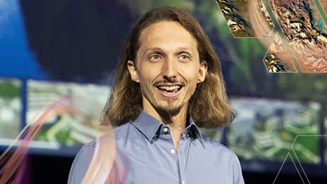 Smiling event presenter wearing a blue dress shirt on stage during a session with mapping graphics in the background