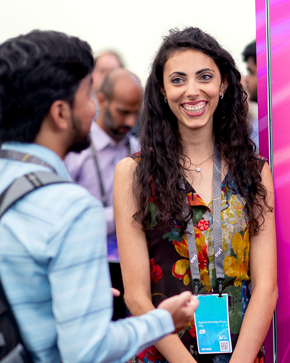 A person wearing a conference lanyard, smiling, and talking to another person