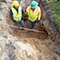 A birds-eye view of two people wearing safety vests and hard hats standing in a deep muddy ditch discussing a protruding pipe