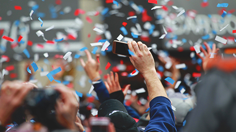 A person in a crowd holding up a camera, recording confetti falling to the ground.