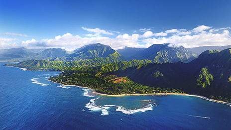 The coastline of Kauai featuring dark blue ocean, vibrant green mountains, and a sky with light clouds.
