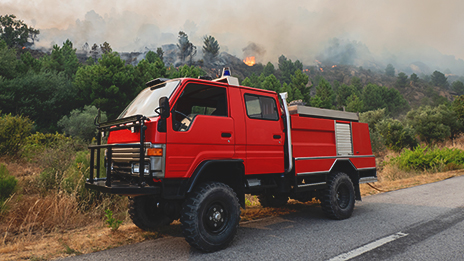 A Portugal fire truck parked outside a forest with a fire in the distance.