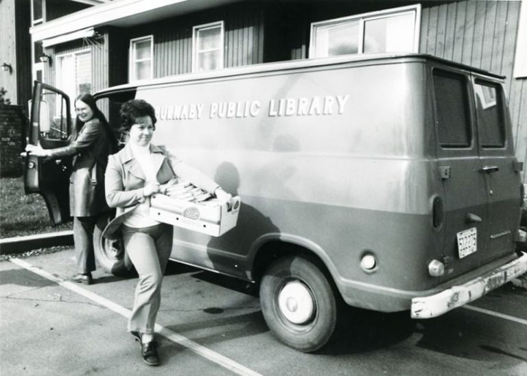 An old, black-and-white photo of a woman carrying a crate of books next to a van labeled Burnaby Public Library