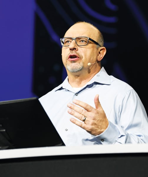 A photo of Jim McKinney talking into a microphone and gesturing with his hands during an onstage demonstration