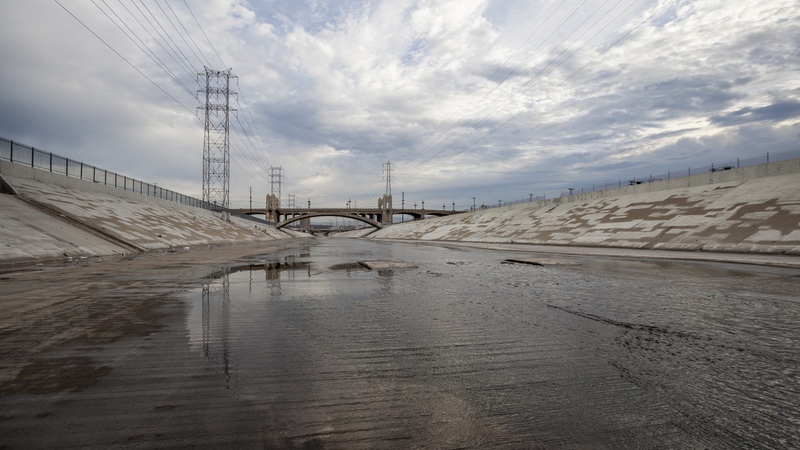 A wide concrete flood control channel with a shallow stream, power line towers, and a bridge in the distance under a cloudy sky.