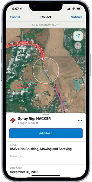 A mobile phone app shows an overhead photo with wooded areas, tan-colored ground, rectangular structures, and a road or path. A circle outline in the middle of the image contains a crosshair symbol.