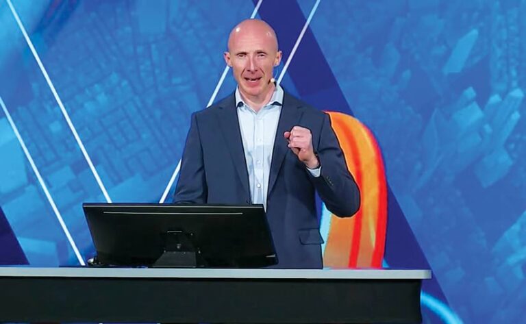 A man, holding his fist in front of his chest to make a point, speaks onstage while standing behind a small computer screen