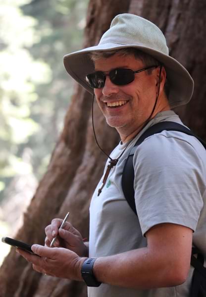 A man wearing a hat, sunglasses, and T-shirt smiles over his shoulder.