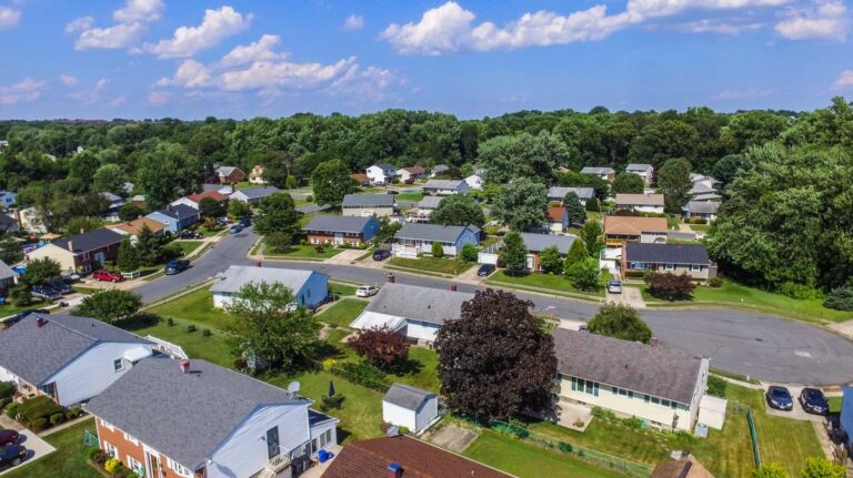 Aerial view of a modest suburban neighborhood with small single-family homes, yards, and tree-lined streets in summer.