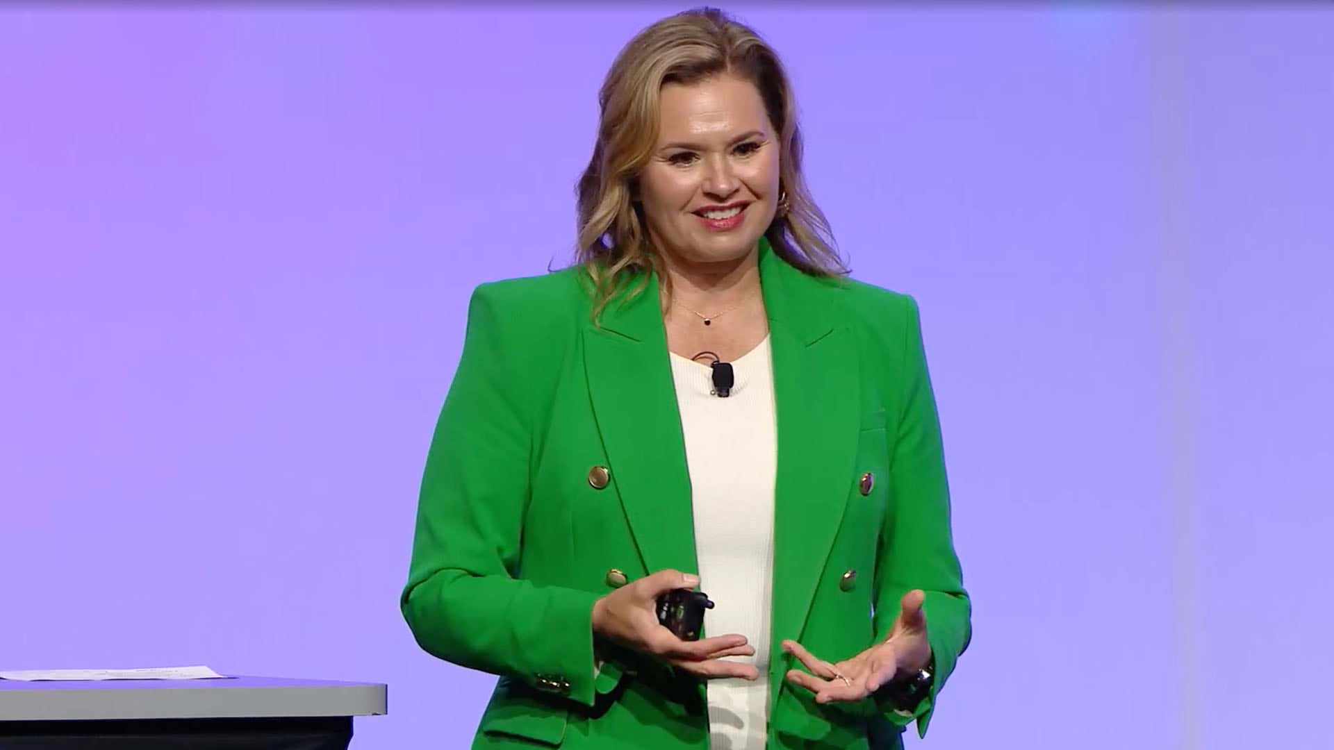A person wearing a green jacket speaking on a stage in front of a lavender background