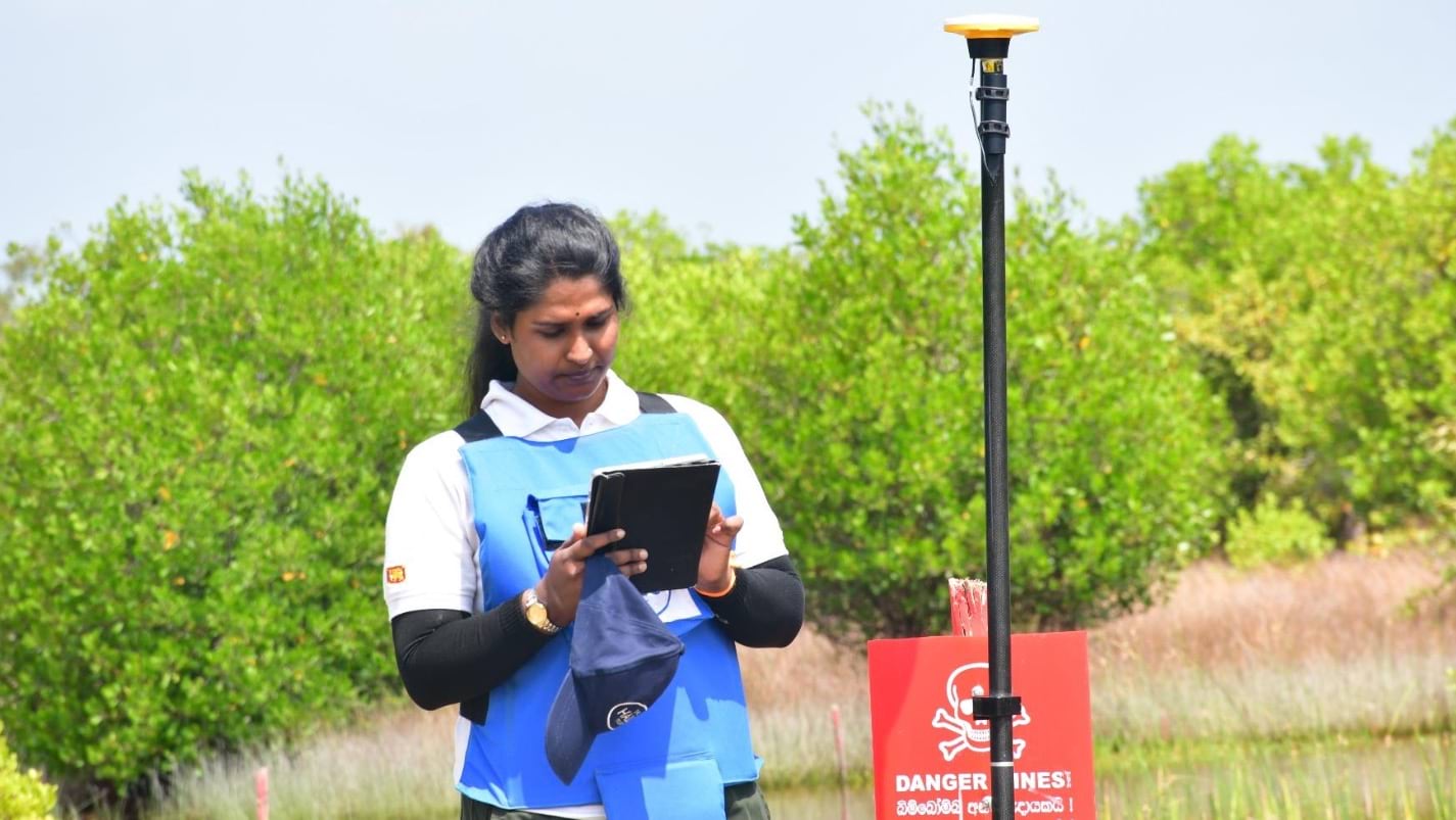 A woman uses a tablet and a GNSS receiver at a demining site