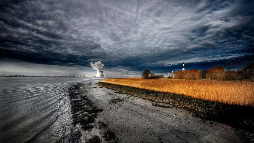 Under a dramatic sky, a power plant sits beyond a black-sand beach with orange beach plants set back from the water