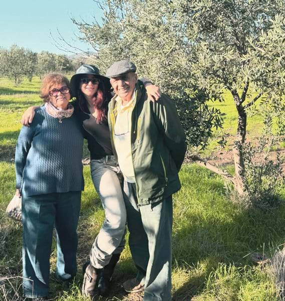 A woman with her arms around her grandparents, against a backdrop of lush olive groves.