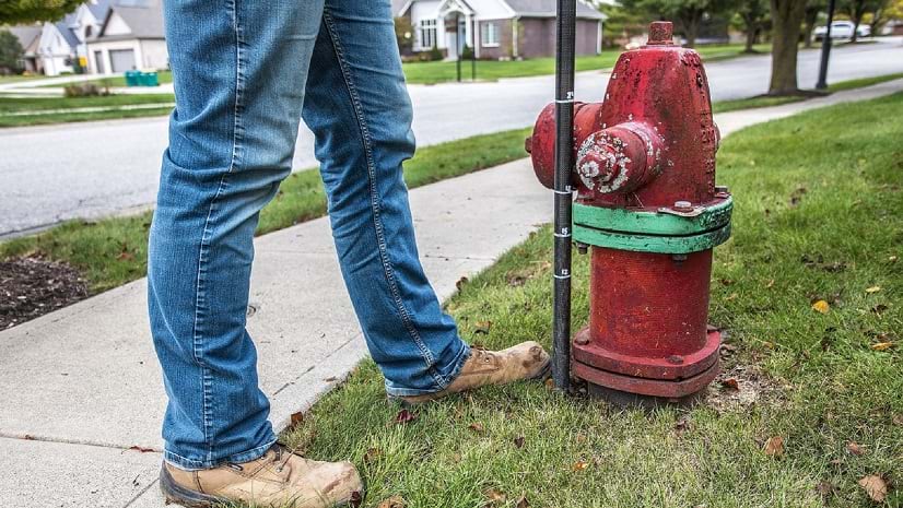 Seen from below the waist, an individual wearing boots and jeans stands next to a measuring pole and a red fire hydrant.