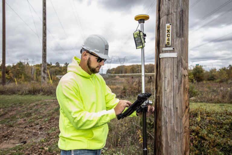 A man wearing a hard hat and a yellow hoodie stands next to a pole and looks at a computer tablet.