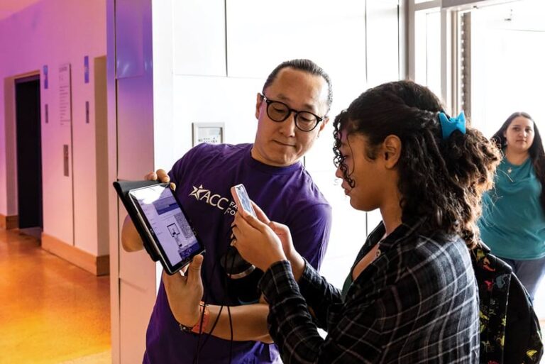 A man holding a tablet up for a college student, who is taking a picture of the tablet display with her phone.