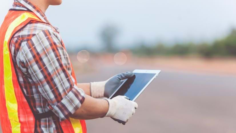 With a paved area in the background, a person wearing an orange and yellow vest and work gloves holds a smart tablet