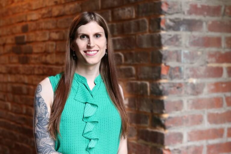 A smiling woman with long brown hair and visible tattoos on her right arm wears a ruffled green sleeveless top. She stands against a backdrop of an exposed brick wall.