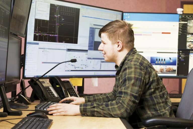 A young man sits at a desk in front of multiple computer screens displaying various data, including maps and graphs. He is wearing a plaid shirt and appears to be working intently. A telephone headset sits on the desk.