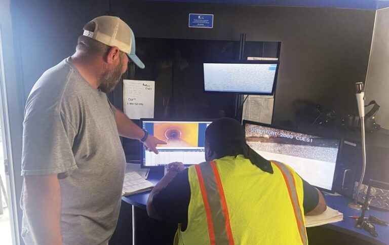 Two men review a computer screen showing an internal pipe inspection. One man, wearing a gray shirt and baseball cap, points to the screen. The other man, wearing a yellow safety vest, looks at the screen. Multiple computer monitors display data and images.