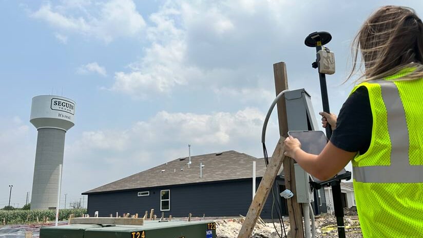 A surveyor uses a tablet and a robotic total station to conduct a land survey at a new home construction site. A water tower reading "Seguin, Texas, It's real" is visible in the background.