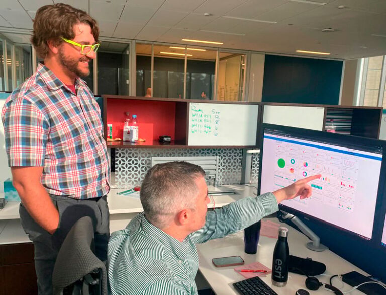 Two men in a modern office are reviewing data on a computer screen. One man, wearing a plaid shirt, points to the screen that displays various charts and graphs. The other man, wearing a button-down shirt, observes. A whiteboard with notes is visible in the background.