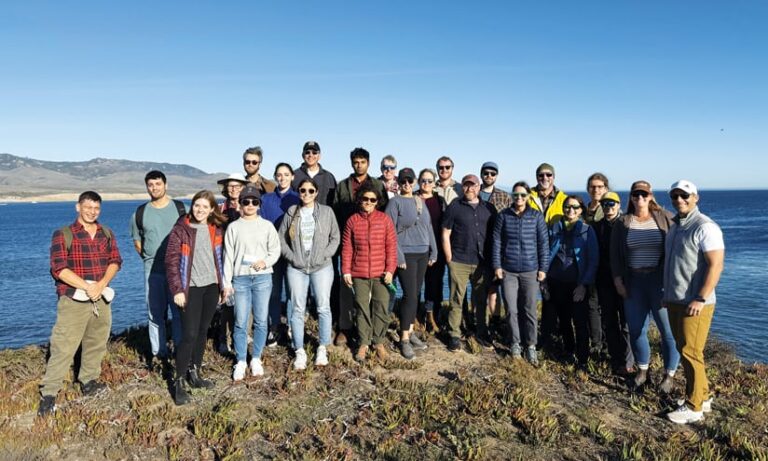 A group of 24 people standing on a bluff with the ocean in the background.