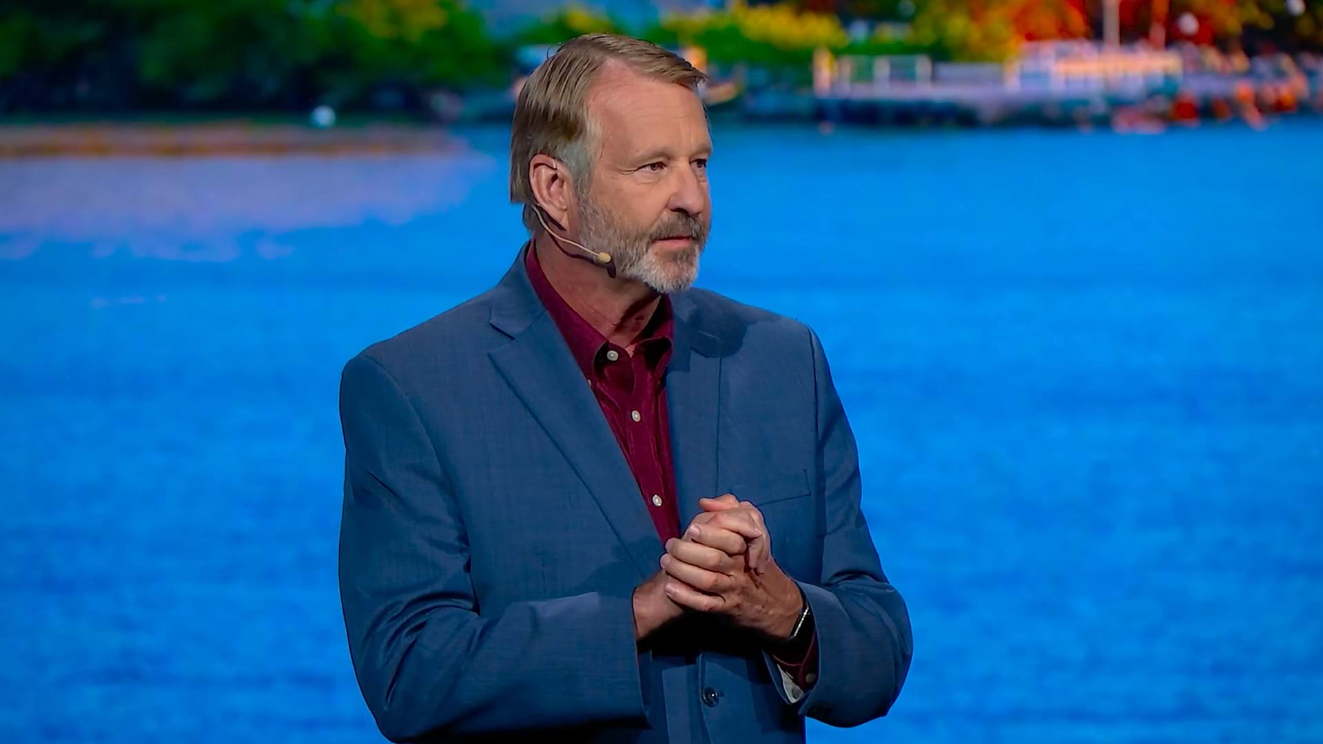 A person wearing a blue suit and clasping their hands in front of a large screen displaying a body of water