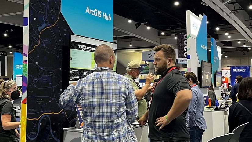 Two people stand in a large room with tables, signs, and other people in the background