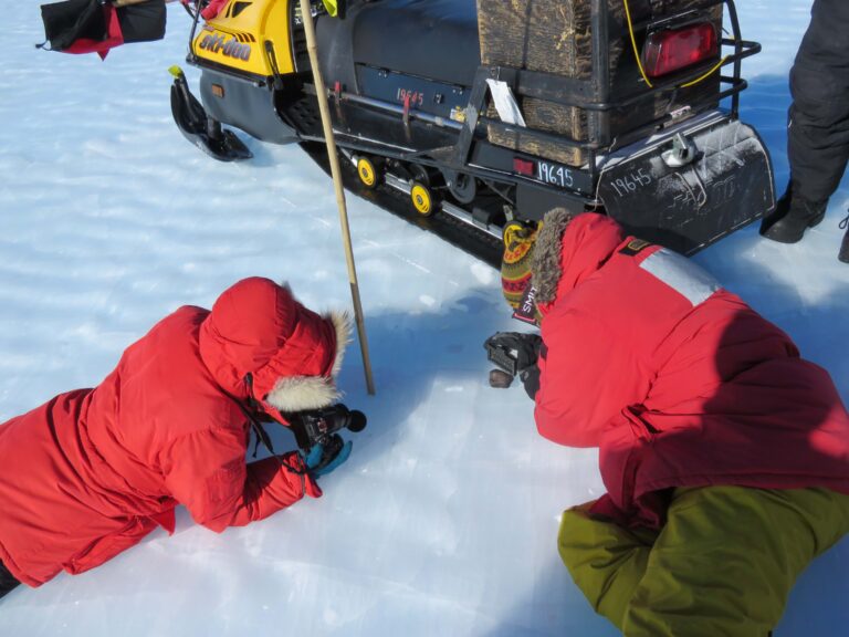 Two people in red parkas on the ground examining snow