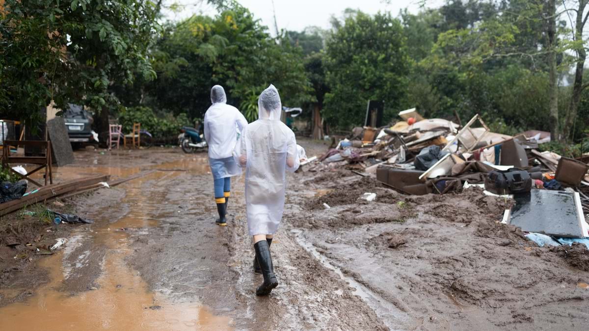 Two workers in white protective suits and boots walking through flood-damaged area with large piles of debris, damaged furniture, and mud covering the ground, with houses and trees visible in the background.