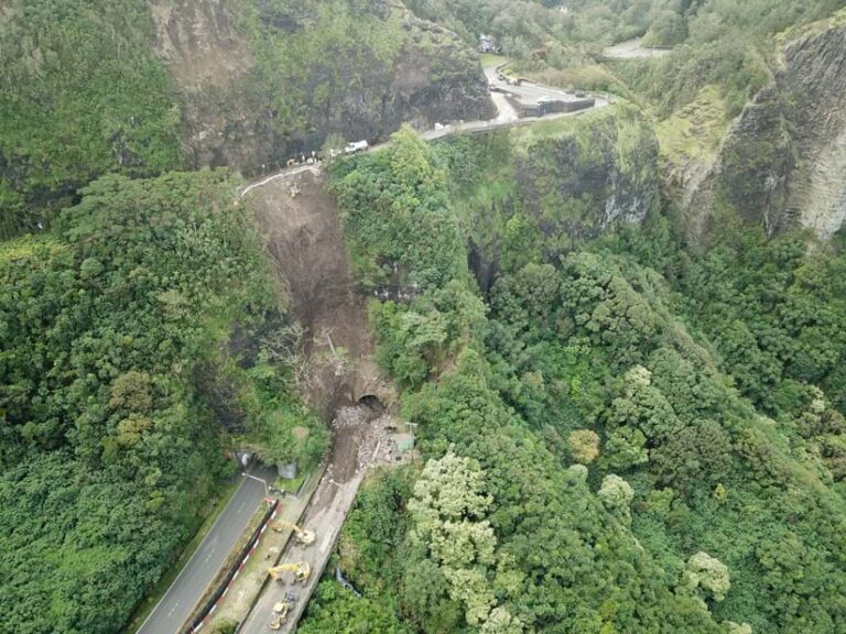 Photo of a highway surrounded by lush vegetation and high cliffs. The highway is blocked by mud and debris from a landslide, while vehicles on the road help to clear the debris.