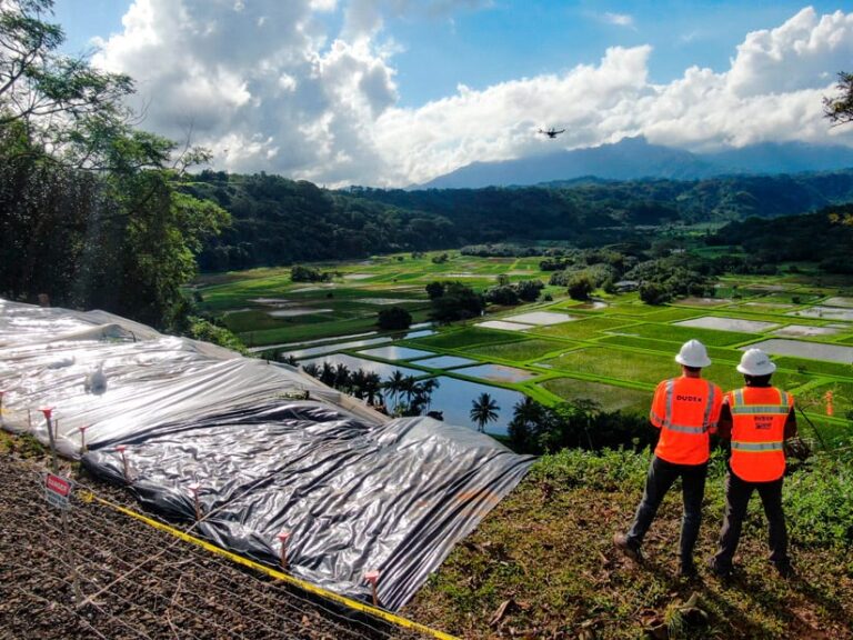 Two men in hardhats and hi-vis vests stand on a hill overlooking a farm. A drone, operated by the men, appears to be capturing imagery of the site.
