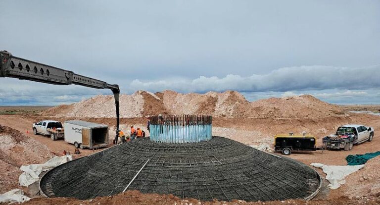 Construction workers and vehicles gathered around a conical structure supported by rebar. Concrete is being poured to form the foundation of a future wind turbine.