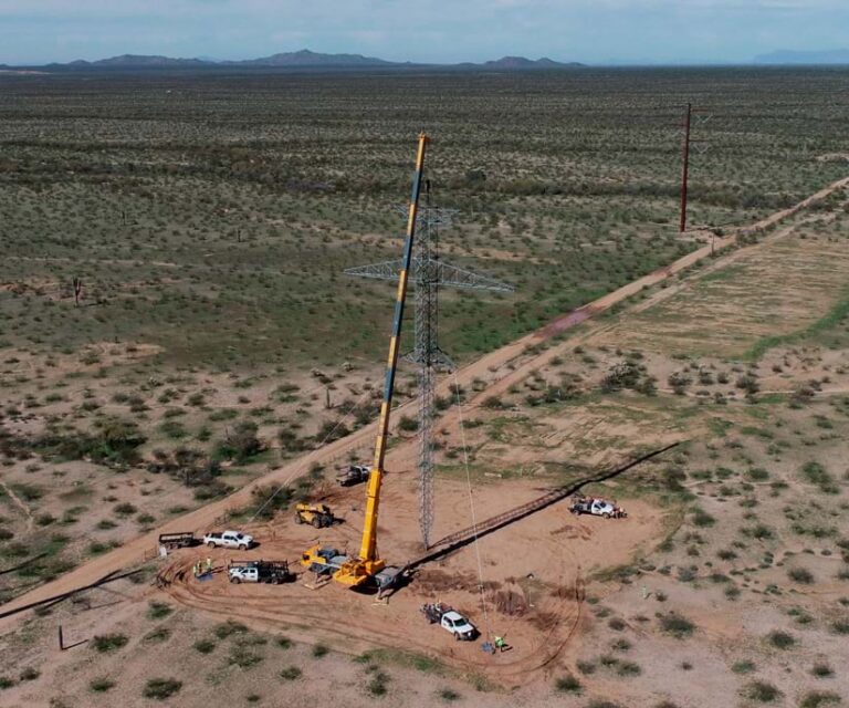 Aerial photograph of a transmission tower under construction, in the middle of a sparse landscape, surrounded by workers and vehicles. The tower is supported by a large crane.