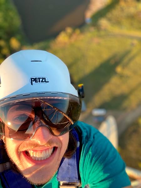 Close-up headshot of a smiling man’s face as he climbs a cell tower, with the ground out of focus in the background.