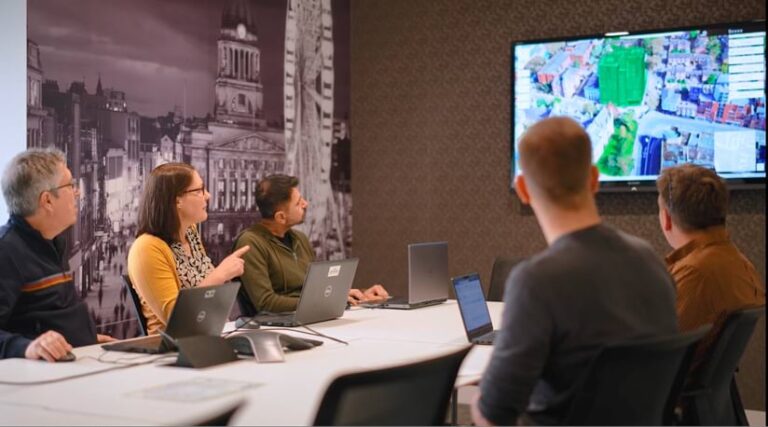 Photograph of five people seated around a conference table, each with a laptop in front of them. The people are looking at a 3D model of a city displayed on a wall-mounted monitor and appear to be discussing it.