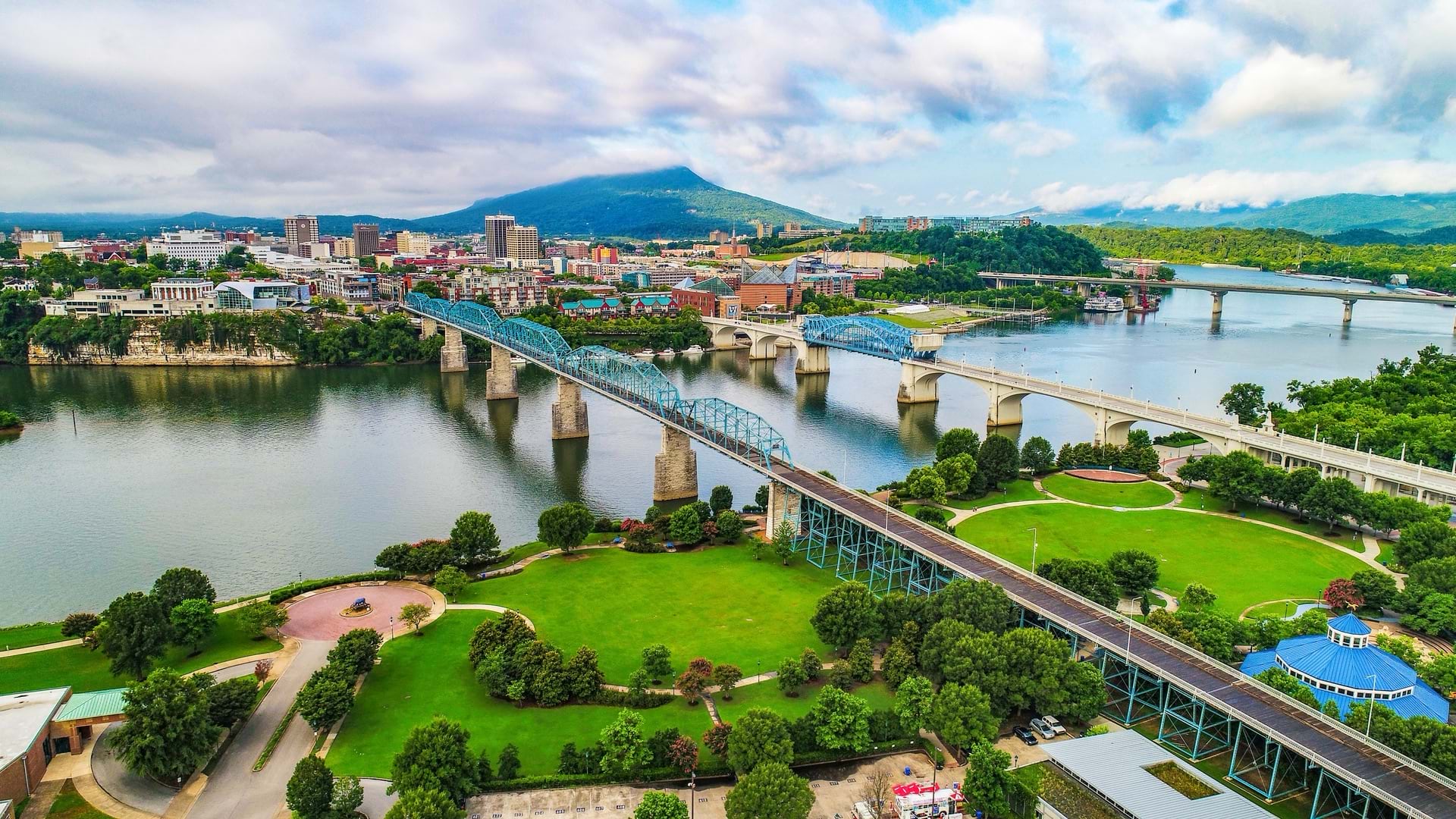 Aerial view of downtown Chattanooga showing the historic Walnut Street Bridge and Market Street Bridge crossing the Tennessee River with expansive green parks along the waterfront, downtown buildings, and Lookout Mountain in the background