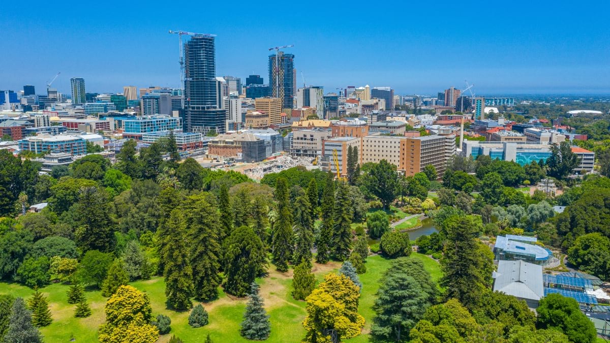 Aerial drone photograph of Adelaide showing the city center skyline surrounded by extensive parklands with green spaces, water features, and scattered trees forming a buffer between urban development and residential areas