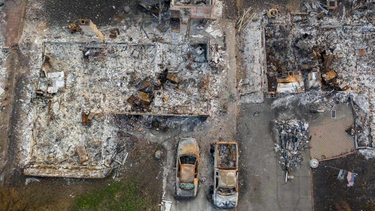 Aerial view of fire-destroyed residential neighborhood showing burned home foundations, charred vehicles in driveways, and ash-covered ground with only concrete slabs and debris remaining where houses once stood.