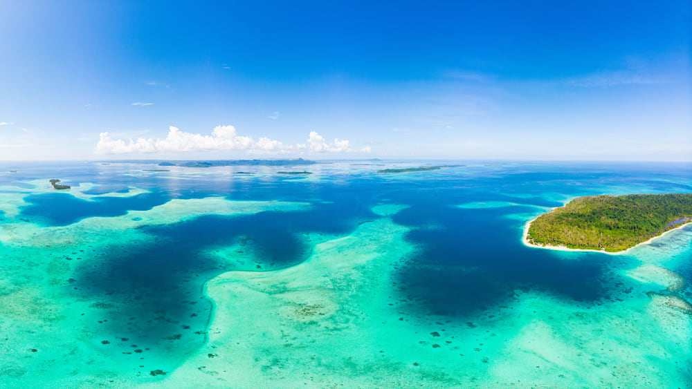 erial view of a tropical atoll showing a forested island surrounded by shallow turquoise lagoons and coral reefs, with deeper blue ocean waters beyond and scattered white clouds against a bright blue sky.