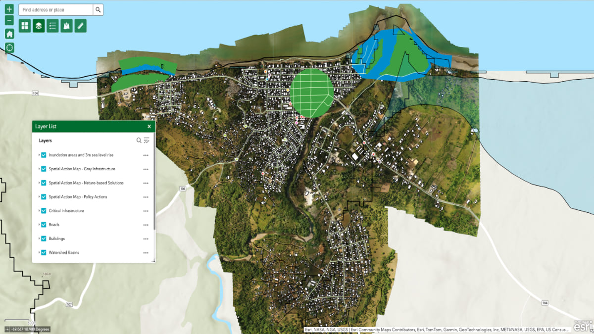 Aerial satellite view of a coastal community with an interactive GIS mapping interface showing various data layers including inundation areas, spatial action maps, infrastructure, roads, buildings, and watershed basins. The map displays urban development patterns alongside green protected areas and blue marine zones, with a layer list panel visible on the left side of the interface.