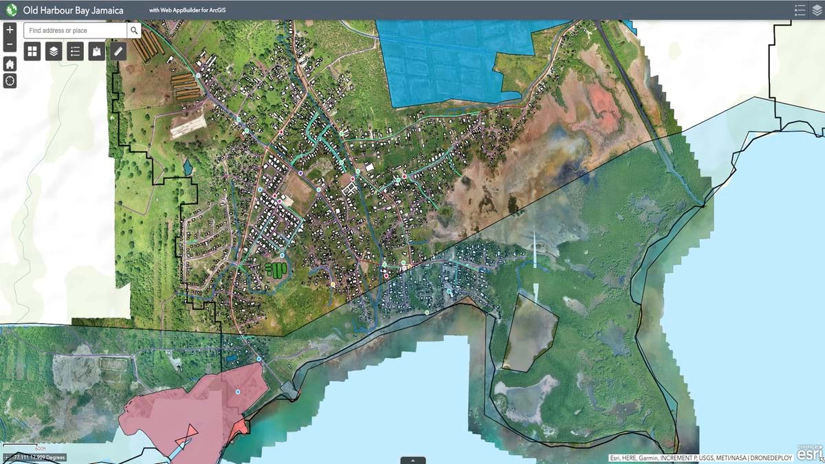Aerial satellite view and GIS map of Old Harbour Bay, Jamaica showing the coastal town's layout with detailed street networks, a prominent pink-shaded area along the southern coastline, green agricultural fields to the north, and the characteristic bay formation with clear turquoise waters. The mapping interface displays various planning layers over the satellite imagery of this Caribbean coastal community.