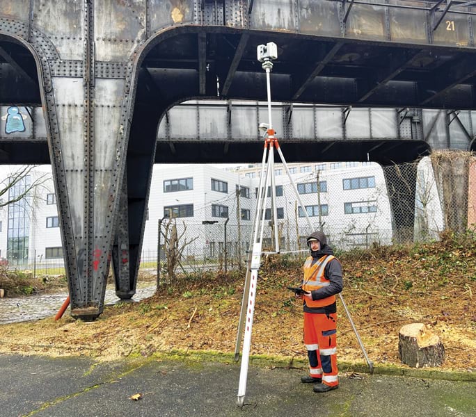 A surveyor in an orange work suit stands near a surveying instrument mounted on a tripod, beneath a large, riveted steel viaduct. A modern building is in the background.