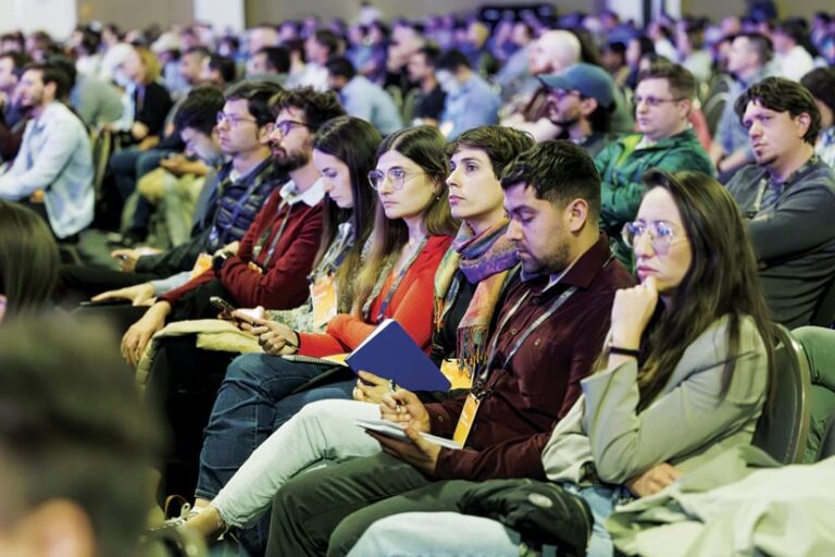 Audience members seated in rows listen attentively during the Esri Developer & Technology Summit Plenary Session. Many are taking notes with pens, phones, and tablets.