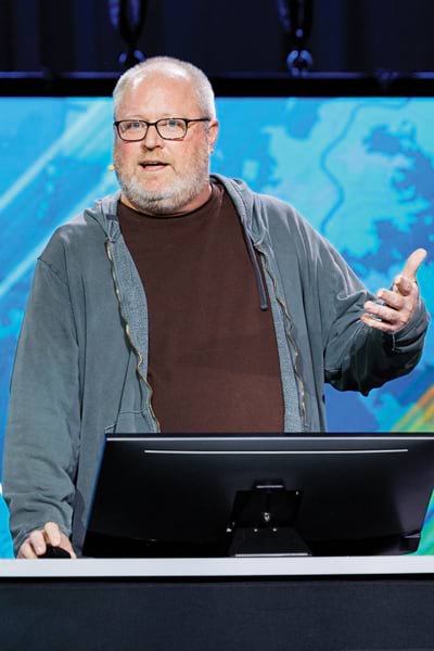 A man wearing glasses, a brown shirt, and a gray zip-up hoodie stands behind a lectern onstage at the Plenary Session gesturing to the audience. Behind him is a screen with a blue, map-like pattern.
