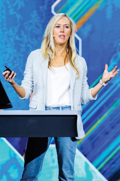 A blonde woman stands on stage behind a podium, holding a clicker and gesturing. She wears jeans, a white top, and a gray blazer. A colorful backdrop is behind her.