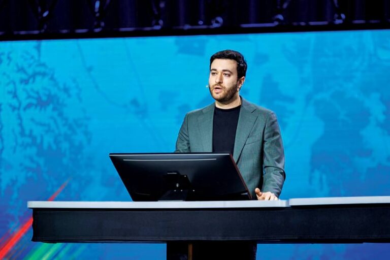 A man with short, dark hair stands behind a podium, speaking. He wears a dark shirt and a gray blazer. The backdrop is a blue screen with abstract patterns.