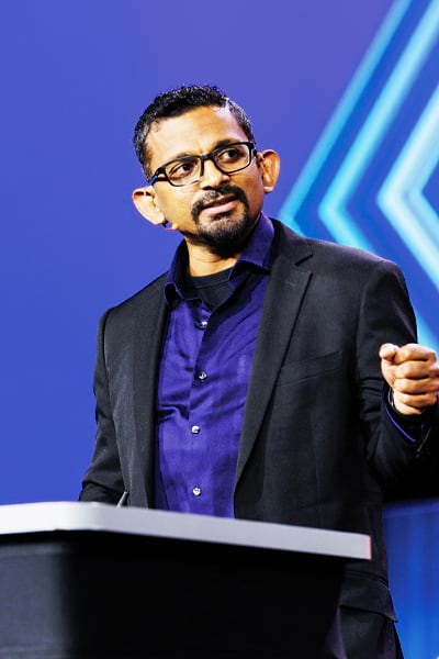 A man with short, dark hair, glasses, and a goatee speaks at a podium. He wears a purple button-down shirt and a black jacket. He is gesturing with his right hand. The background is bright blue.